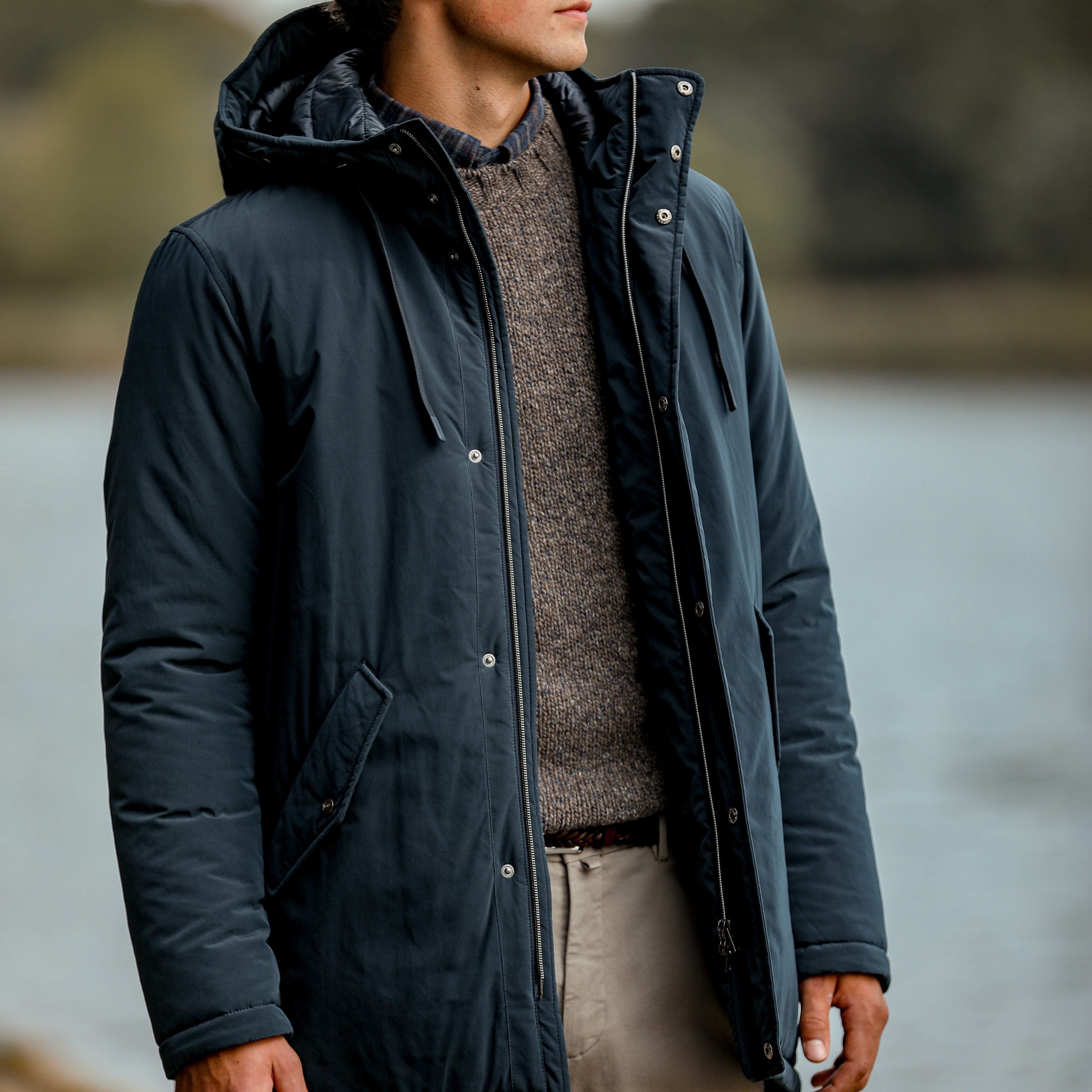 A person stands outdoors near the water, wearing a Herno Dark Blue Nylon Technical Padded Jacket over a sweater and light-colored pants.