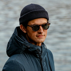 A person wearing a beanie, a jacket, and The Bespoke Dudes' Welt Earth Bio Gradient Bottle Lenses 46mm sunglasses with small rounded frames stands outdoors near a body of water.