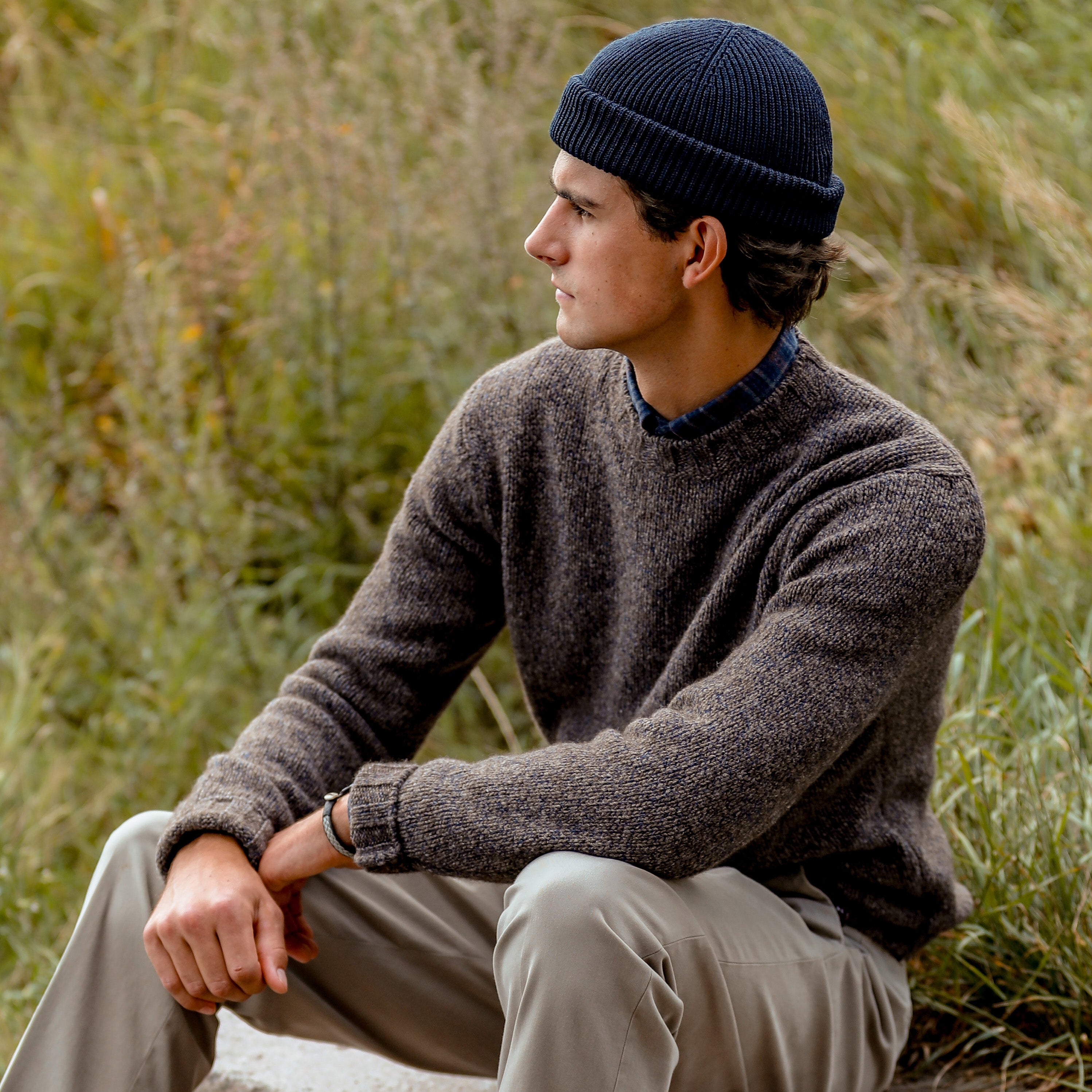 A person wearing a dark beanie, a Brown Melange Yak Cashmere Sweater by Filippo de Laurentiis, and beige pants sits outdoors on a grassy area, looking to the side.