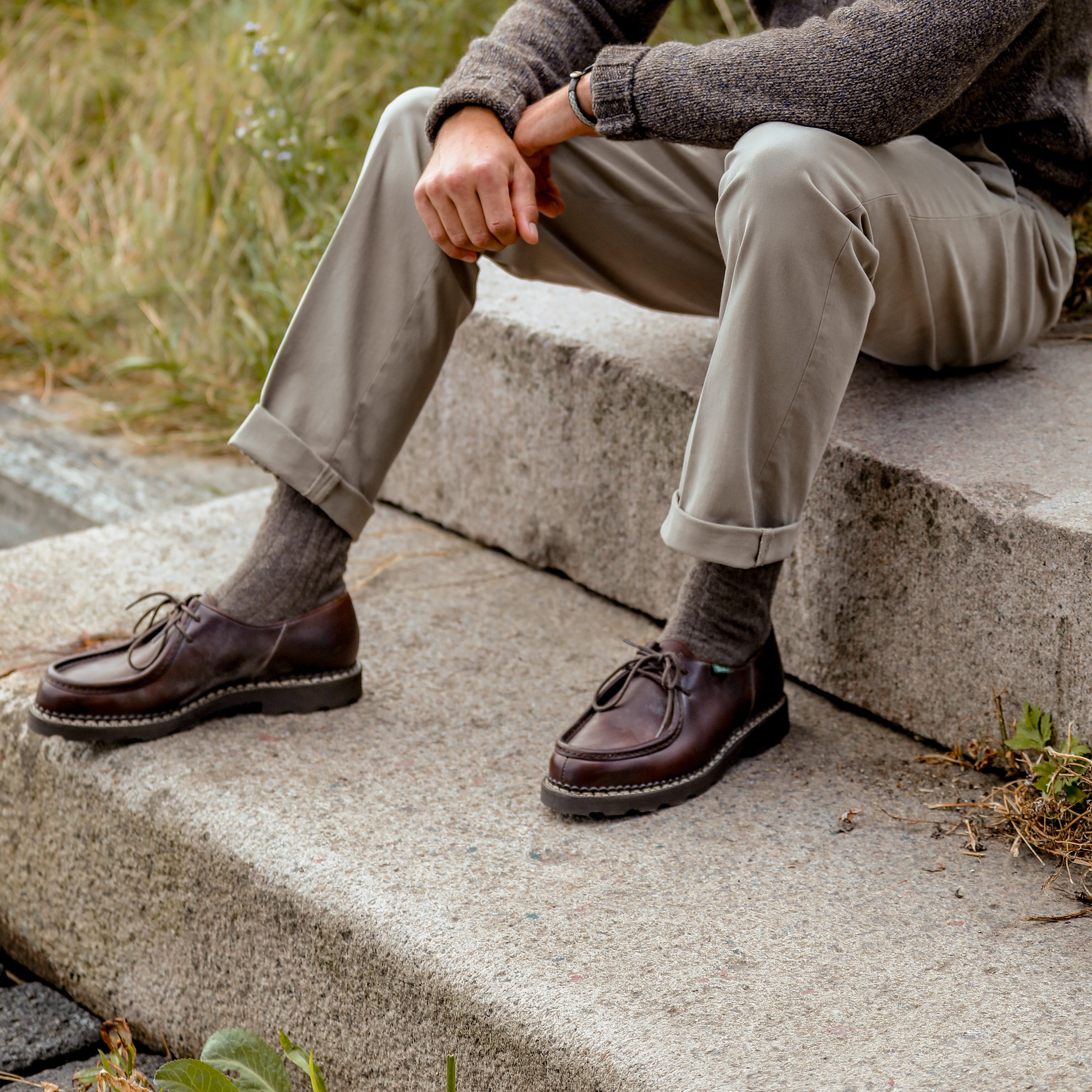 A person wearing beige pants, a gray sweater, and brown Lis Cafe Leather Michael Derbies from Paraboot sits on stone steps outdoors.