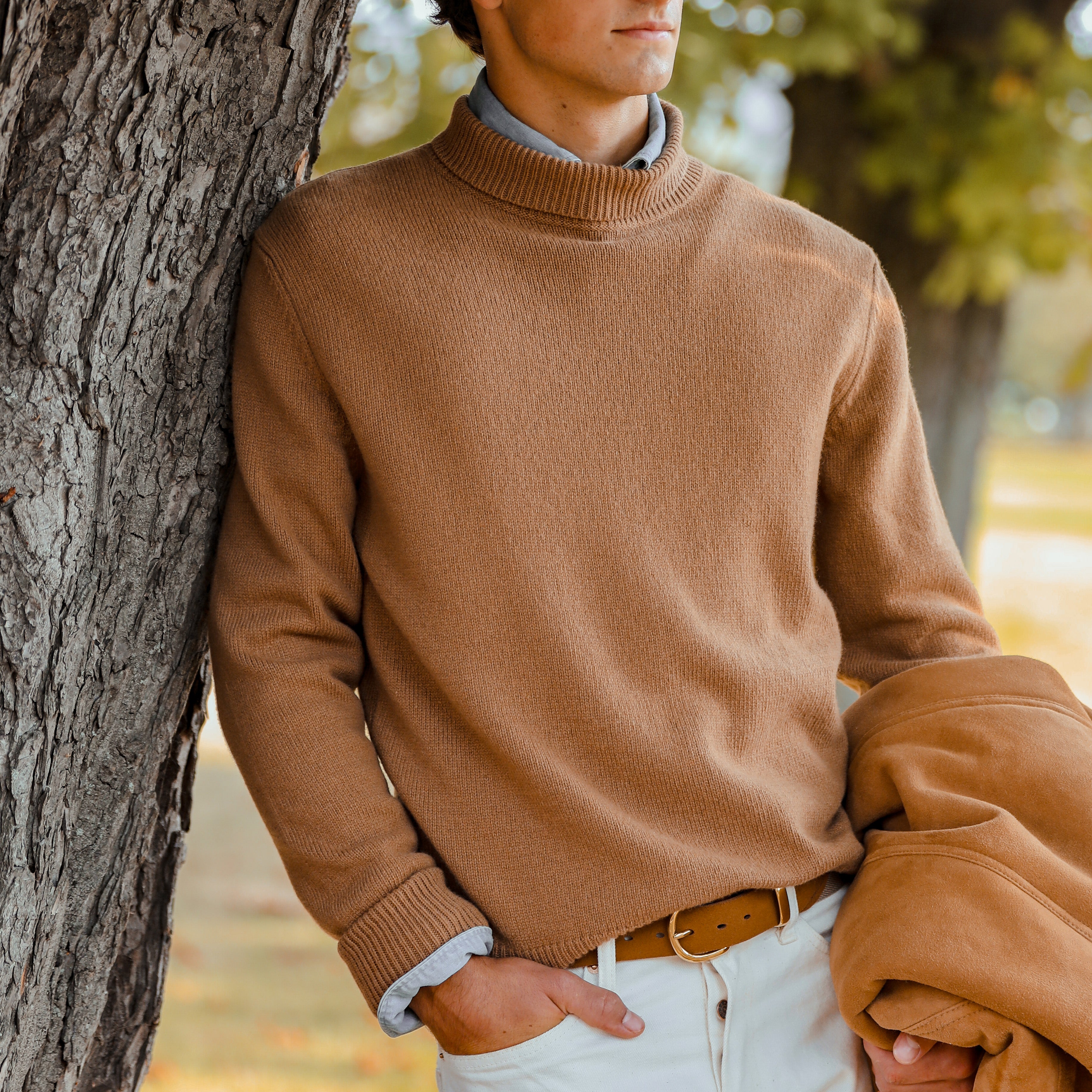 A person in a contemporary fit, Camel Beige Wool Cashmere Mock Neck Sweater by G.R.P stands outdoors, leaning against a tree. They hold a wool and cashmere brown coat over their arm and wear white pants.