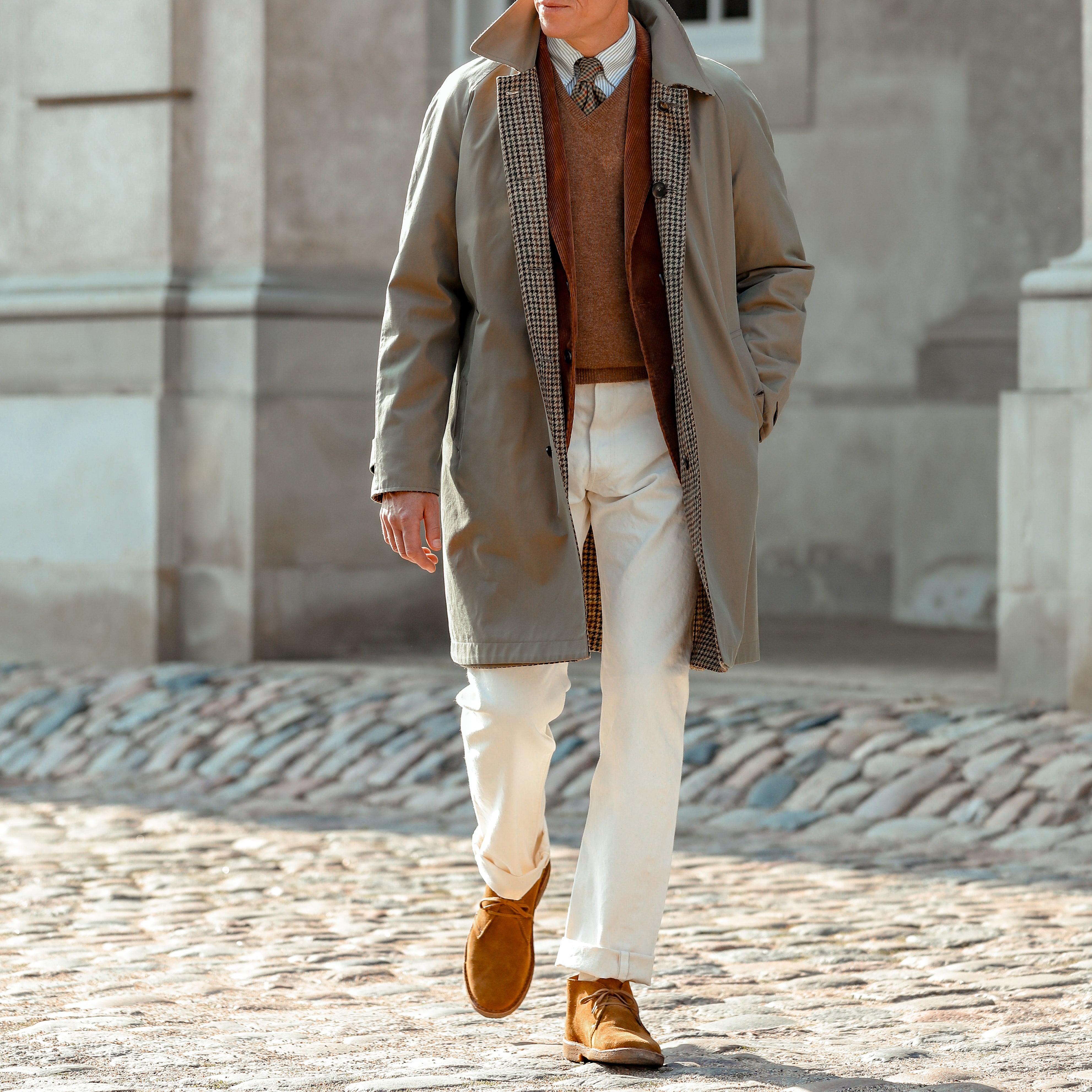 A person walks outdoors wearing the Beige Houndstooth Wool Tweed Reversible Coat by L'Impermeabile, a rust-brown sweater, white pants, tan suede shoes, and a tie. The background is an urban stone building.