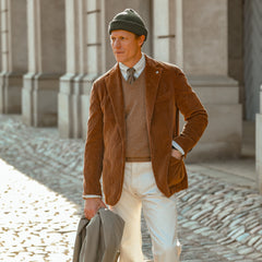 A man in an L.B.M. 1911 Cinnamon Brown Cotton Corduroy Blazer, white pants, and a green knit beanie holds a light-colored coat over cobblestone pavement in front of a building.