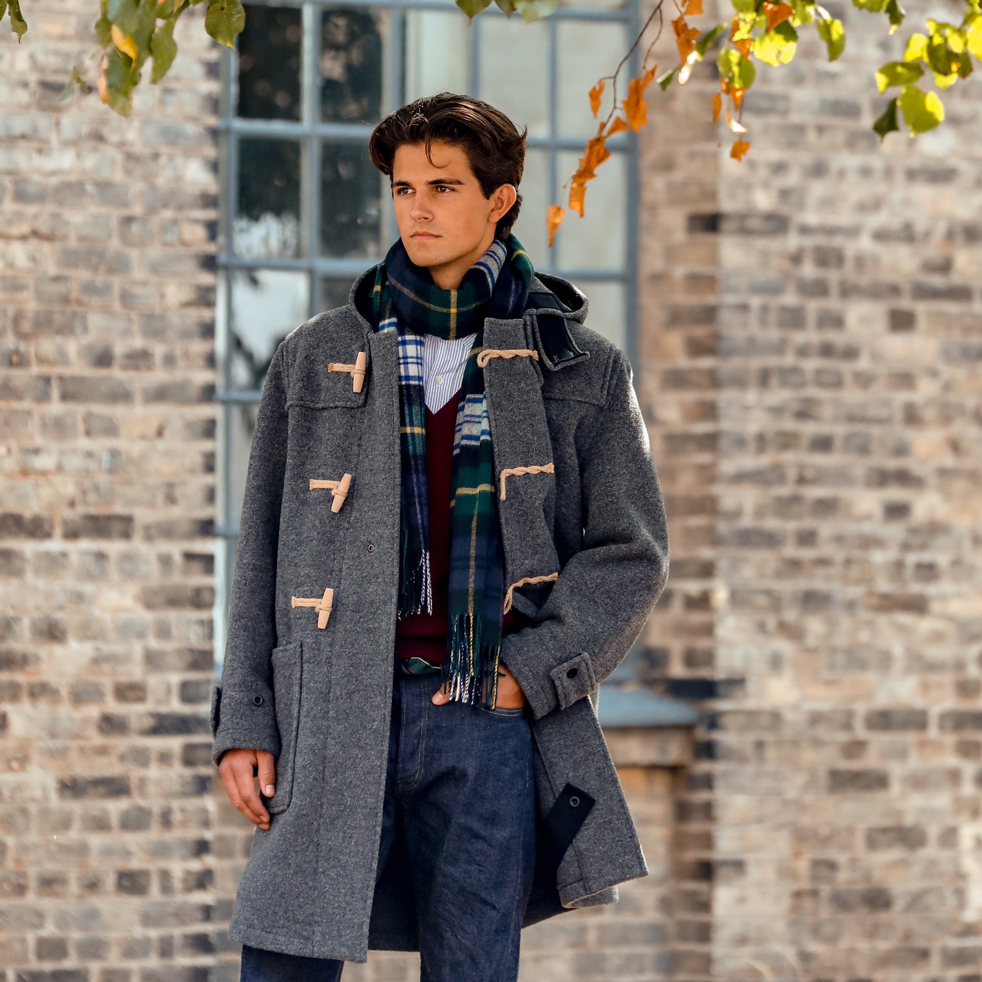 A man stands outdoors wearing the Gloverall Grey Melange Wool Monty Duffle Coat with toggle buttons, a colorful scarf, and blue pants. He is in front of a brick building with windows.