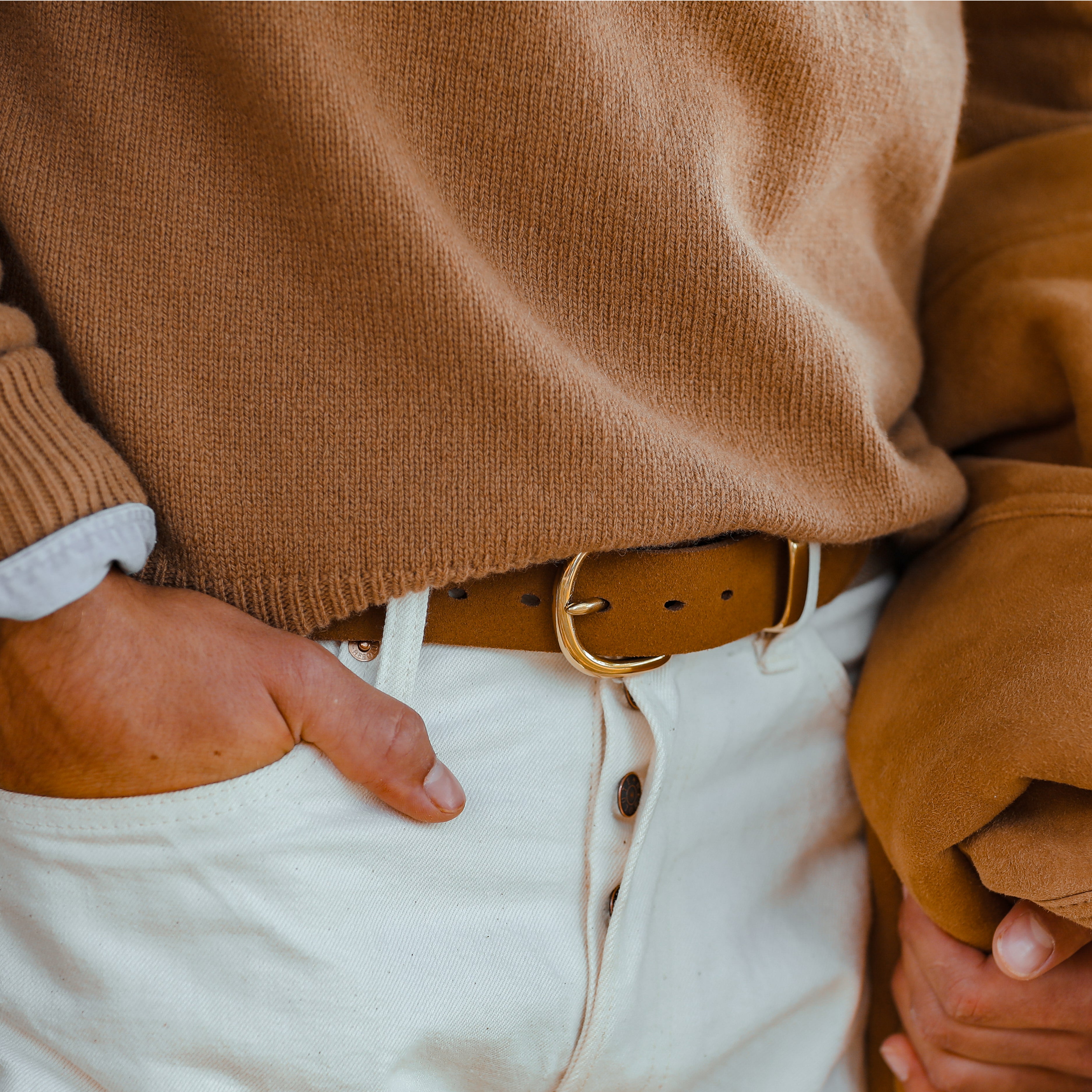 A person wearing a tan sweater and white pants with their right hand in their pocket. The person is also holding a tan jacket, accentuated by the Beige Brown Striped Canvas Cognac Suede 35mm Belt from Hardy & Parsons.