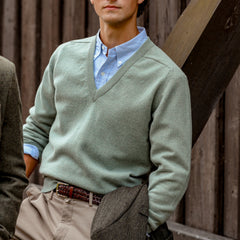 A person is wearing a Mojito Green Deep V-Neck Lambswool Sweater by William Lockie over a blue collared shirt and khaki pants, holding a brown coat. They are standing in front of a wooden wall with diagonal beams.