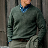 A person wearing a William Lockie Spruce Green Shetland Lambswool V-Neck Sweater over a denim shirt stands against a wooden background, holding a supersoft lambswool jacket.