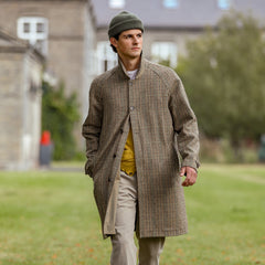 A man wearing a L'Impermeabile Beige Houndstooth Wool Tweed Reversible Coat, a yellow vest, beige pants, and a green beanie is walking outdoors on a lawn with buildings in the background.