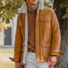 A person wearing a Werner Christ Camel Beige Suede Leather Liam Jacket with fur lining over a brown sweater and white pants stands outdoors near a tree.