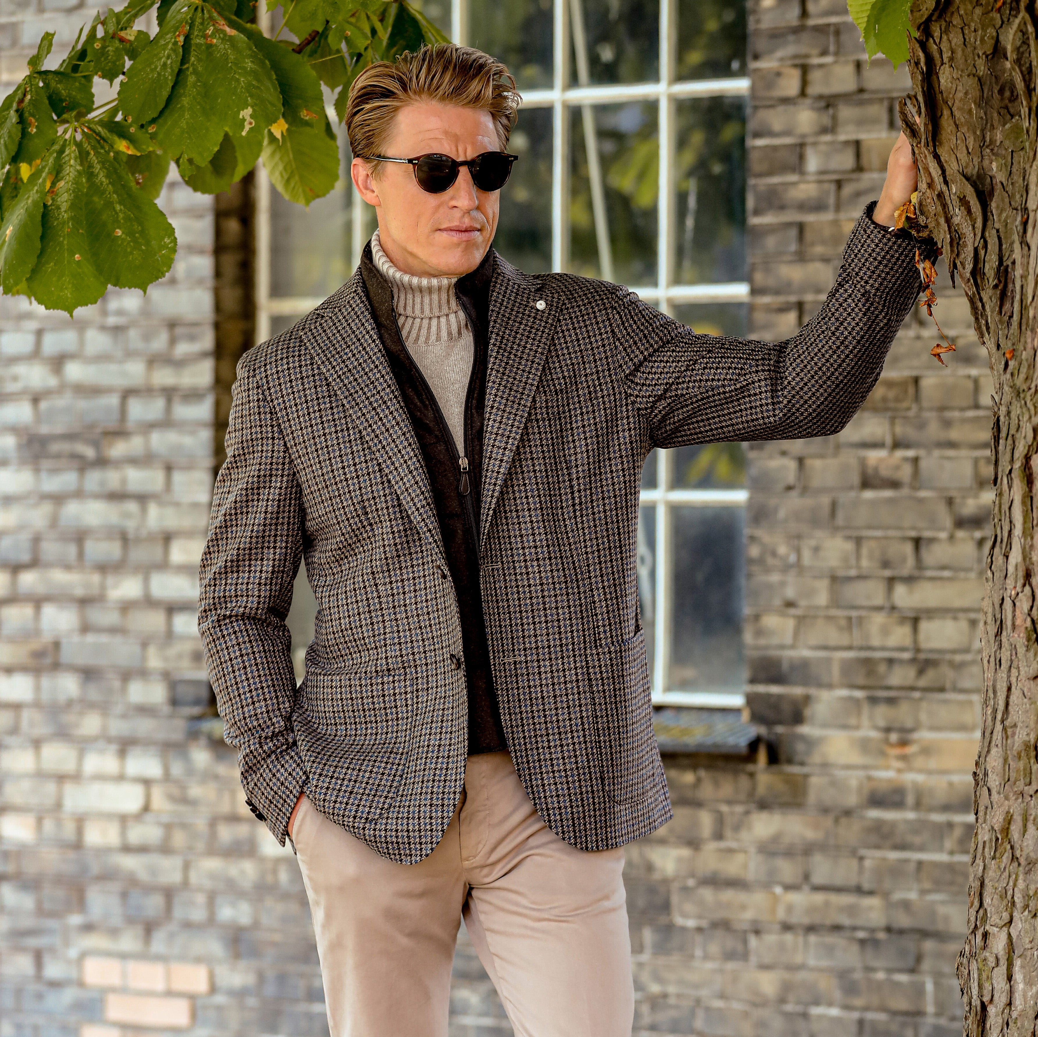 A man wearing sunglasses, a Luigi Bianchi Brown Houndstooth Wool Cashmere Hybrid Blazer with horn buttons, and a turtleneck sweater stands outdoors near a tree, leaning against it with one hand.