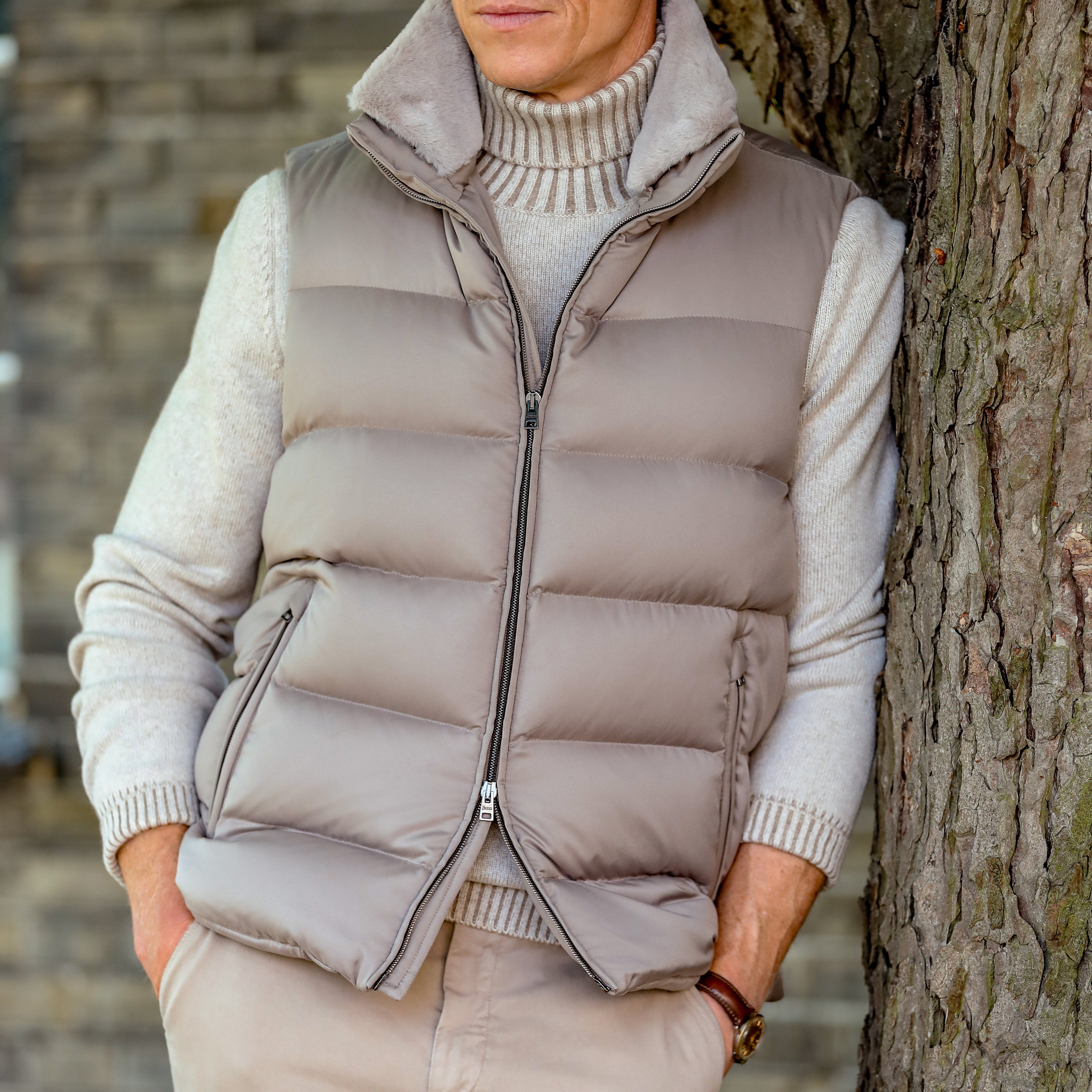 A person wearing a Light Beige Nylon Goose Down Quilted Gilet by Herno over a light gray turtleneck sweater leans against a tree.