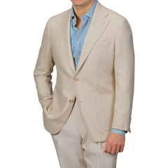 A man wearing a Baltzar Sartorial Beige Wool Silk Summer Tweed Blazer with a blue dress shirt poses with one hand in his pocket against a plain gray background.