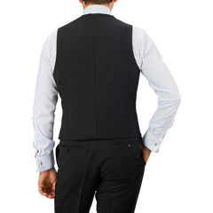 A person in a Baltzar Sartorial Black Wool Mohair Tuxedo Waistcoat and white dress shirt is seen from the back, with hands resting on hips.