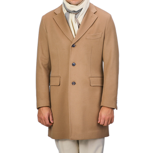 A person is dressed in a Baltzar Sartorial tailored coat named Camel Beige Loro Piana, crafted from pure wool and featuring Rain System technology, along with a white scarf and beige pants. The face is not visible.