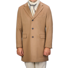 A person is dressed in a Baltzar Sartorial tailored coat named Camel Beige Loro Piana, crafted from pure wool and featuring Rain System technology, along with a white scarf and beige pants. The face is not visible.