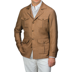 A man wearing a Baltzar Sartorial Camel Brown Wool Silk Linen Safari Jacket with four pockets and white pants stands against a neutral background, effortlessly showcasing a stylish tailored blazer substitute.