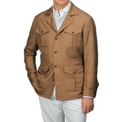 A man wearing a Baltzar Sartorial Camel Brown Wool Silk Linen Safari Jacket with four pockets and white pants stands against a neutral background, effortlessly showcasing a stylish tailored blazer substitute.