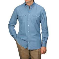 A man stands against a gray background, wearing a Denim Blue Cotton Twill BD Western Shirt by Baltzar Sartorial, paired with brown pants.