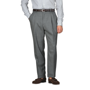 A person is dressed in Baltzar Sartorial Grey High-Twist Wool Pleated Trousers and a striped shirt, hands in pockets, complemented by a brown belt and shoes.