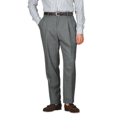 A person is dressed in Baltzar Sartorial Grey High-Twist Wool Pleated Trousers and a striped shirt, hands in pockets, complemented by a brown belt and shoes.