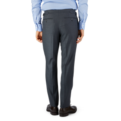 A man wearing a blue shirt and Grey Super 100's Wool Flat Front Suit Trousers by Baltzar Sartorial, standing on a black surface against a blue and gray background.