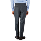 Man standing back-to-camera wearing a blue shirt and Baltzar Sartorial Grey Super 100's Wool Pleated Suit Trousers with black shoes on a plain background.