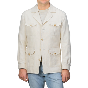 A man in a Light Beige Pure Linen Safari Jacket by Baltzar Sartorial, featuring multiple pockets and mid-weight linen, stands beside light blue jeans against a plain backdrop.