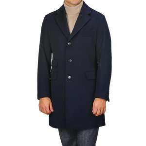 A person is dressed in a Baltzar Sartorial Navy Blue Loro Piana Wool Tailored Coat, showcasing a single-breasted design with three buttons and flap pockets. Underneath, they wear a beige turtleneck sweater paired with gray pants, making it an ideal outfit for any weather due to the innovative Rain System.