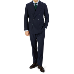 A man in a tailored Navy Blue Super 100s Wool DB Suit Jacket by Baltzar Sartorial, white shirt, and green tie, standing against a gray and white background.