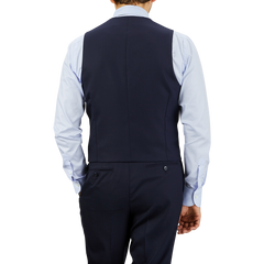 A man viewed from behind wearing a Baltzar Sartorial Navy Super 100s Wool DB Waistcoat and matching trousers with a light blue shirt, standing against a gray background.