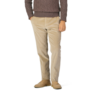 A person wearing a brown cable-knit sweater, Berwich Beige Cotton Corduroy Flat Front Trousers, and tan suede shoes stands with hands in pockets against a plain background.