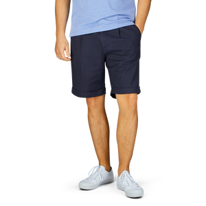 A person wearing a light blue shirt, Berwich navy blue cotton blend pleated shorts with a wider fit, and white sneakers stands against a plain background.