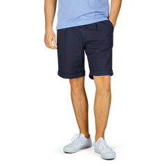 A person wearing a light blue shirt, Berwich navy blue cotton blend pleated shorts with a wider fit, and white sneakers stands against a plain background.