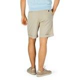 Man standing in Sabbia Beige Cotton Blend Pleated Shorts by Berwich and light-colored sneakers against a blue background.