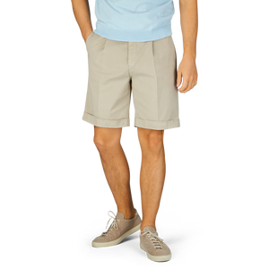 A person wearing Berwich Sabbia Beige Cotton Blend Pleated Shorts and casual shoes against a blue background.