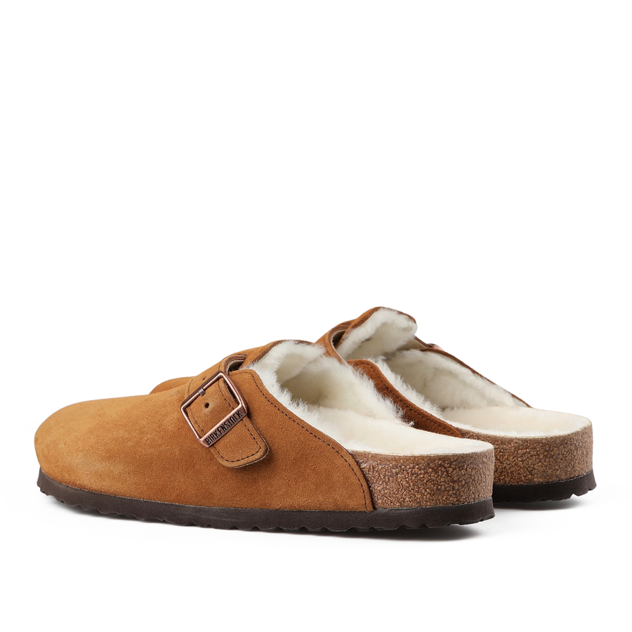 The Mink Brown Suede Shearling Lined Boston Slippers by Birkenstock are a pair of slip-on slippers made from brown suede leather, adorned with buckles and featuring a plush shearling fur lining. They also come with cork soles that echo the classic Birkenstock design.