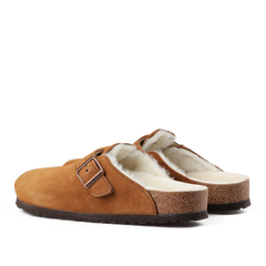 The Mink Brown Suede Shearling Lined Boston Slippers by Birkenstock are a pair of slip-on slippers made from brown suede leather, adorned with buckles and featuring a plush shearling fur lining. They also come with cork soles that echo the classic Birkenstock design.