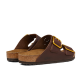 A pair of brown, open-toe sandals with cork soles and two wide straps featuring large gold buckles. The Birkenstock Roast Bold Natural Leather Arizona Sandals, crafted with a natural leather upper, are viewed from the back and side and include an anatomically-supported footbed for added comfort.