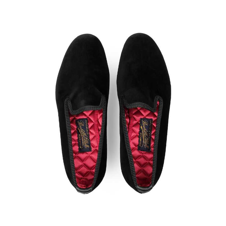 A pair of Plain Black Velvet Albert Slippers by Bowhill Elliott, featuring a luxurious red quilted inner lining and a branded insole logo, designed for style and comfort, viewed from above.