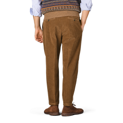 A person stands facing away, wearing Briglia’s Tobacco Pleated Corduroy Courmayeur Trousers, a patterned knit sweater, blue shirt, and brown shoes, against a plain background.