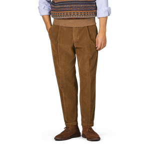 Person wearing Briglia Tobacco Pleated Corduroy Courmayeur Trousers, a blue shirt with rolled-up sleeves, and a sweater vest with a patterned design. Hands in pockets. Only the lower half of the body is visible.