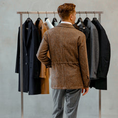 A man wearing a Brown Herringbone Wool Tweed Utility Jacket from Baltzar Sartorial stands facing a clothing rack with various jackets, including other stylish utility options, hanging in a neutral-toned room.