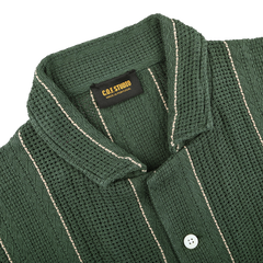 The Dark Green Striped Crochet Relaxed Camp Shirt by C.O.F Studio is made from pure cotton mesh, featuring a camp collar with buttons and white vertical stripes, offering comfort and style. A label inside reads "C.O.F. Studio Made in Portugal.