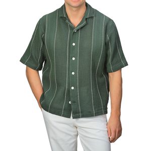 A person stands against a neutral background wearing C.O.F Studio's Dark Green Striped Crochet Relaxed Camp Shirt paired with white pants.