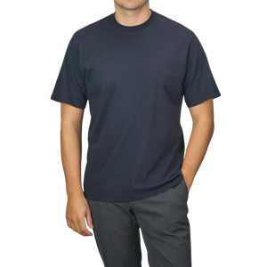 A person wearing a Navy Blue Organic Cotton Crewneck T-Shirt from C.O.F Studio and dark pants stands with one hand in their pocket against a plain background.