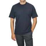 A person wearing a Navy Blue Organic Cotton Crewneck T-Shirt from C.O.F Studio and dark pants stands with one hand in their pocket against a plain background.