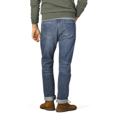 Person standing and facing away, wearing a green long-sleeve shirt, brown shoes, and rolled-up Washed Blue 14oz Kuroki Selvedge Cotton M8 Jeans from C.O.F Studio.