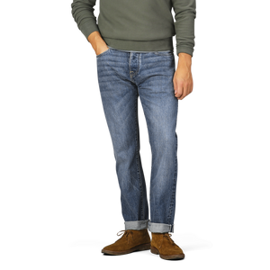 A person wearing a green long-sleeve shirt, C.O.F Studio Washed Blue 14oz Kuroki Selvedge Cotton M8 Jeans with the bottom cuffs rolled up, and brown shoes. Only the lower half of the body is visible.