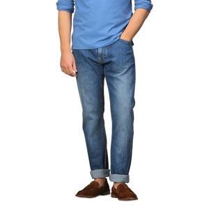 A person in a blue long-sleeve shirt, C.O.F Studio Indigo Blue Washed Organic Cotton M8 Jeans with cuffs, and brown loafers stands against a plain background.