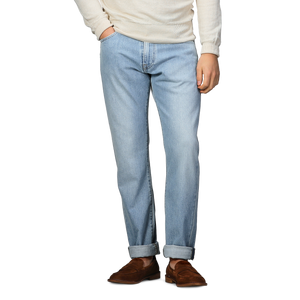 A person is wearing C.O.F Studio's Light Blue 90's Wash Organic Cotton M8 Jeans, featuring a straight roomy fit with rolled cuffs. The look includes a beige long-sleeve shirt and brown shoes, with hands in pockets.